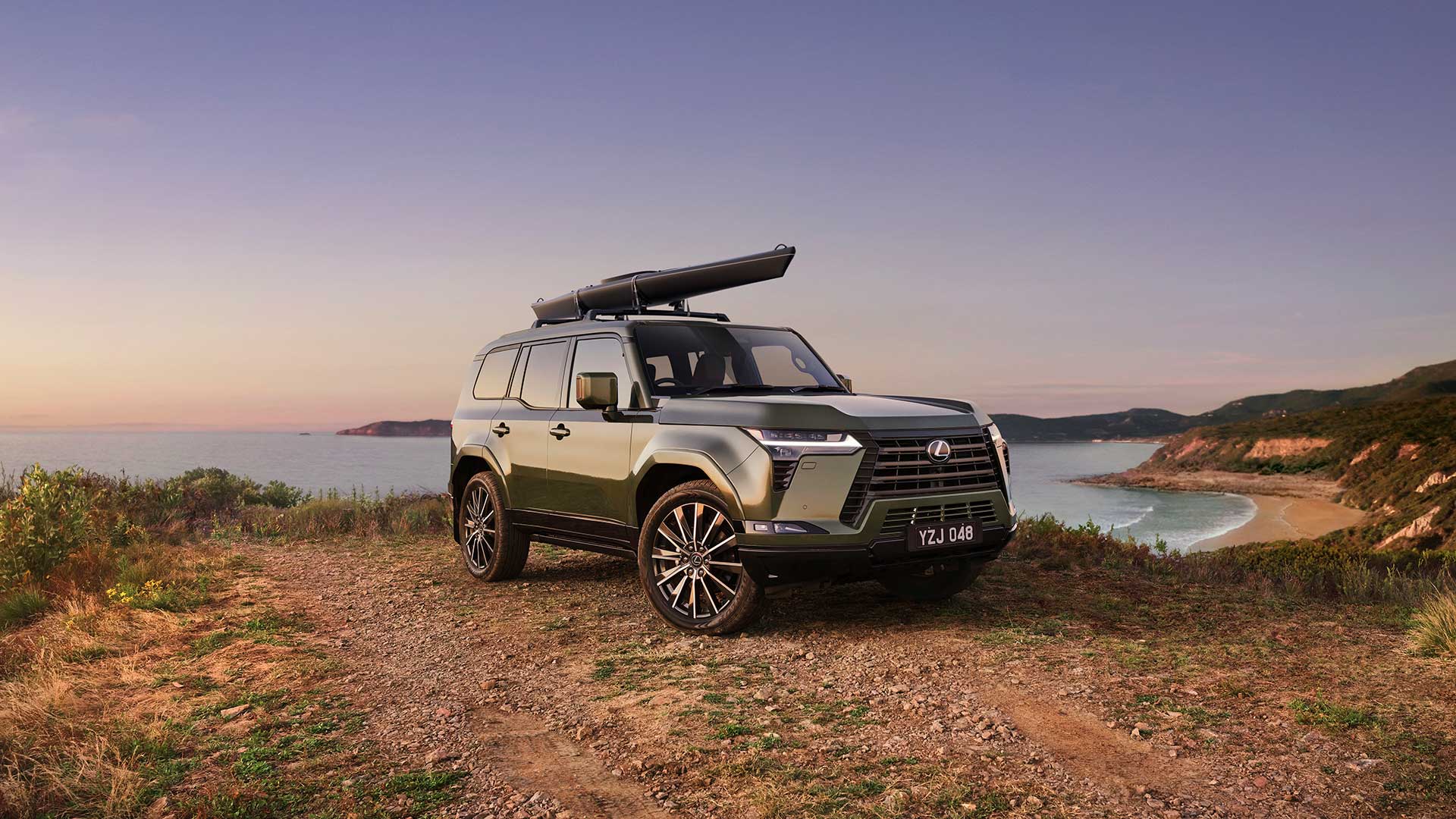 Lexus GX shown parked off-road with optional kayak attached to the roof using optional kayak carrier accessory. The coast is shown in the background. 